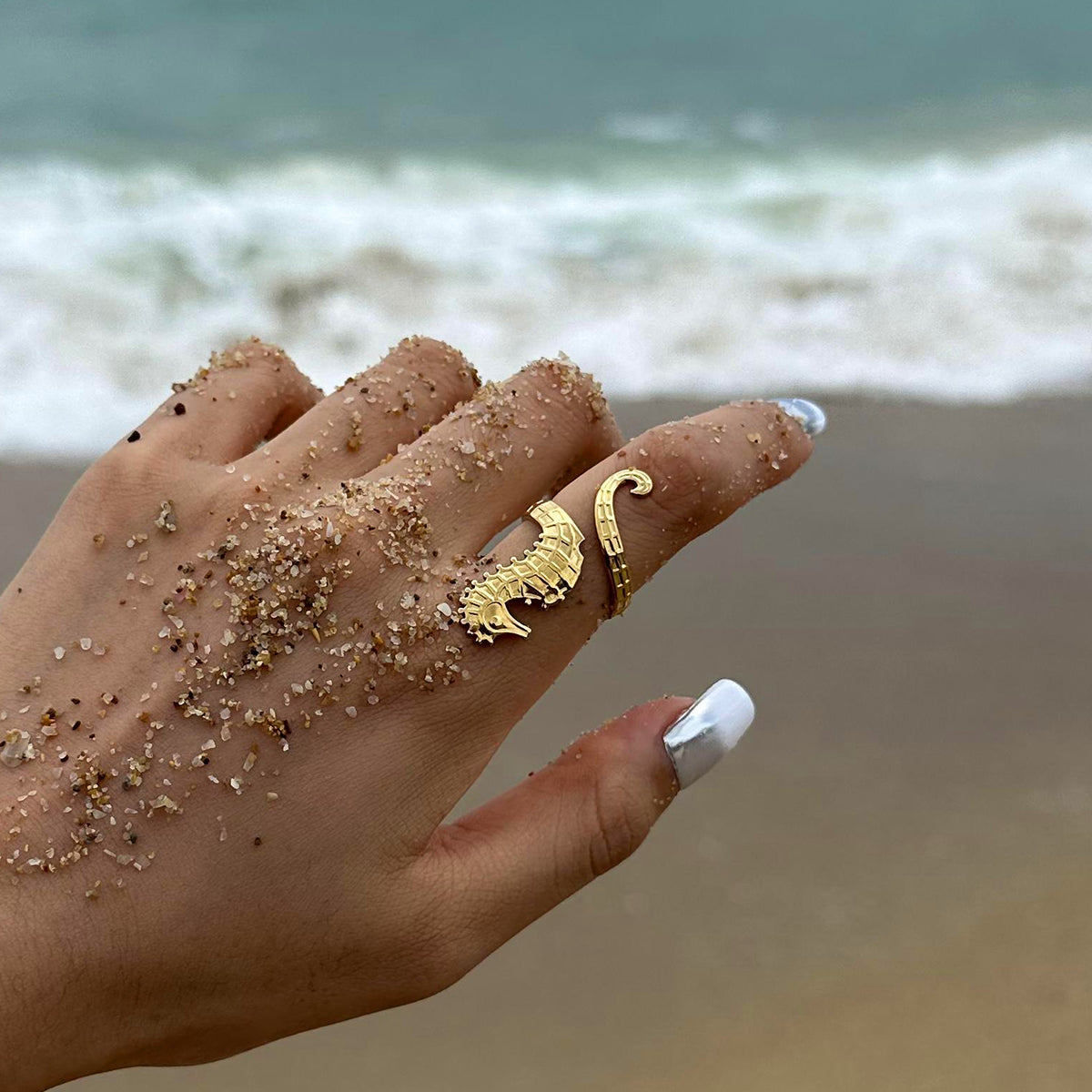 Hand with sand on fingers wearing a gold ring, against a beach background